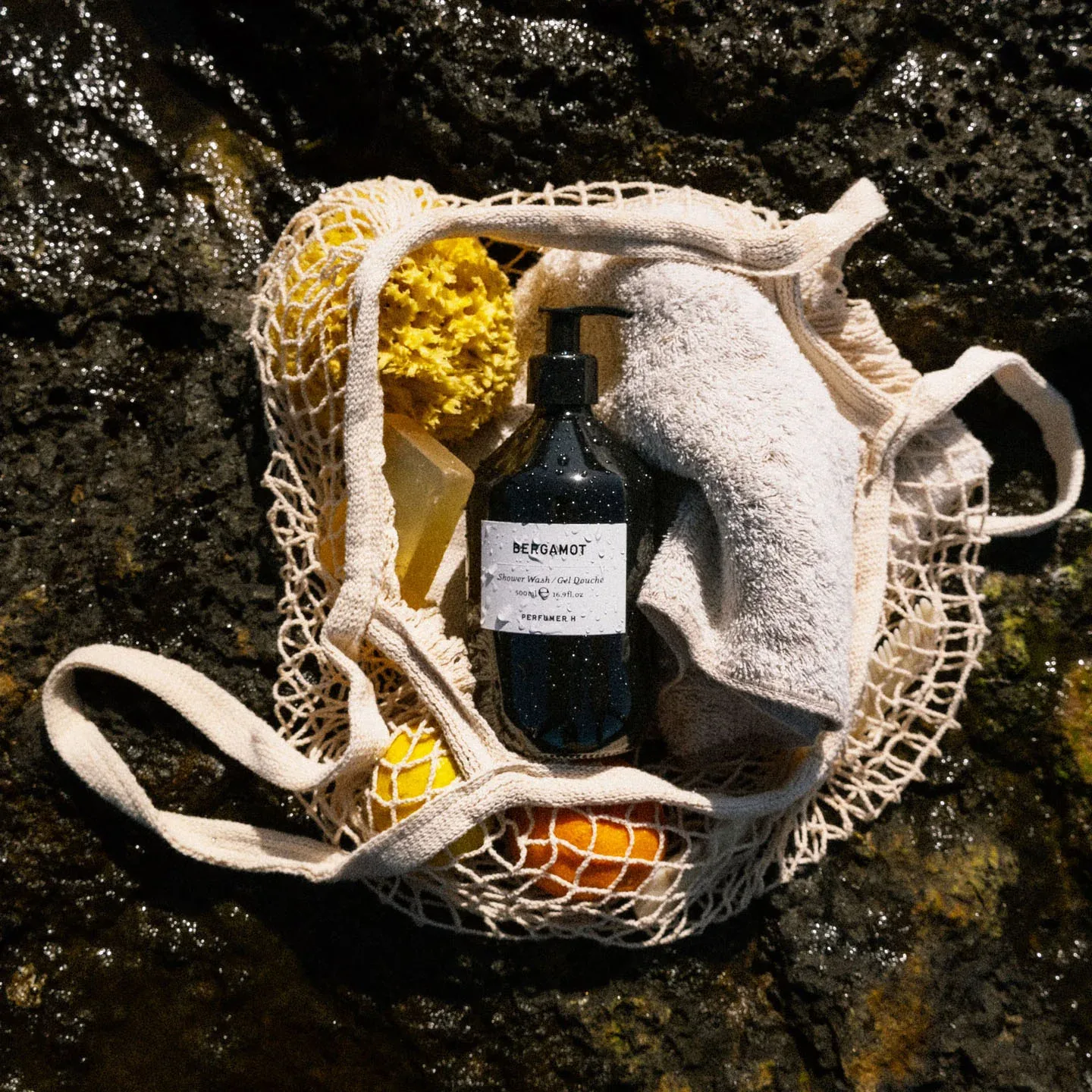 Perfumer H 沐浴瓶放在網袋裡，與柑橘一起置於海岸岩石